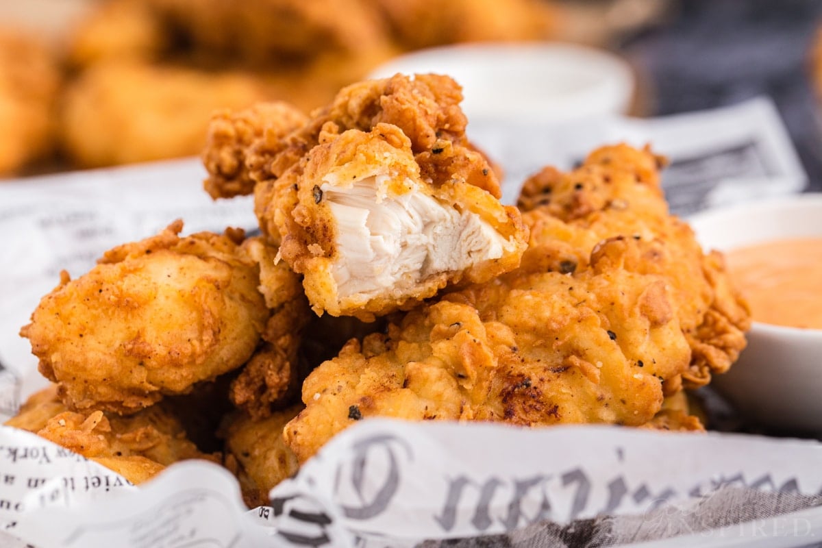 Close-up of buttermilk fried chicken tenders on newspaper wrapping with a bite removed from one of the cutlets.