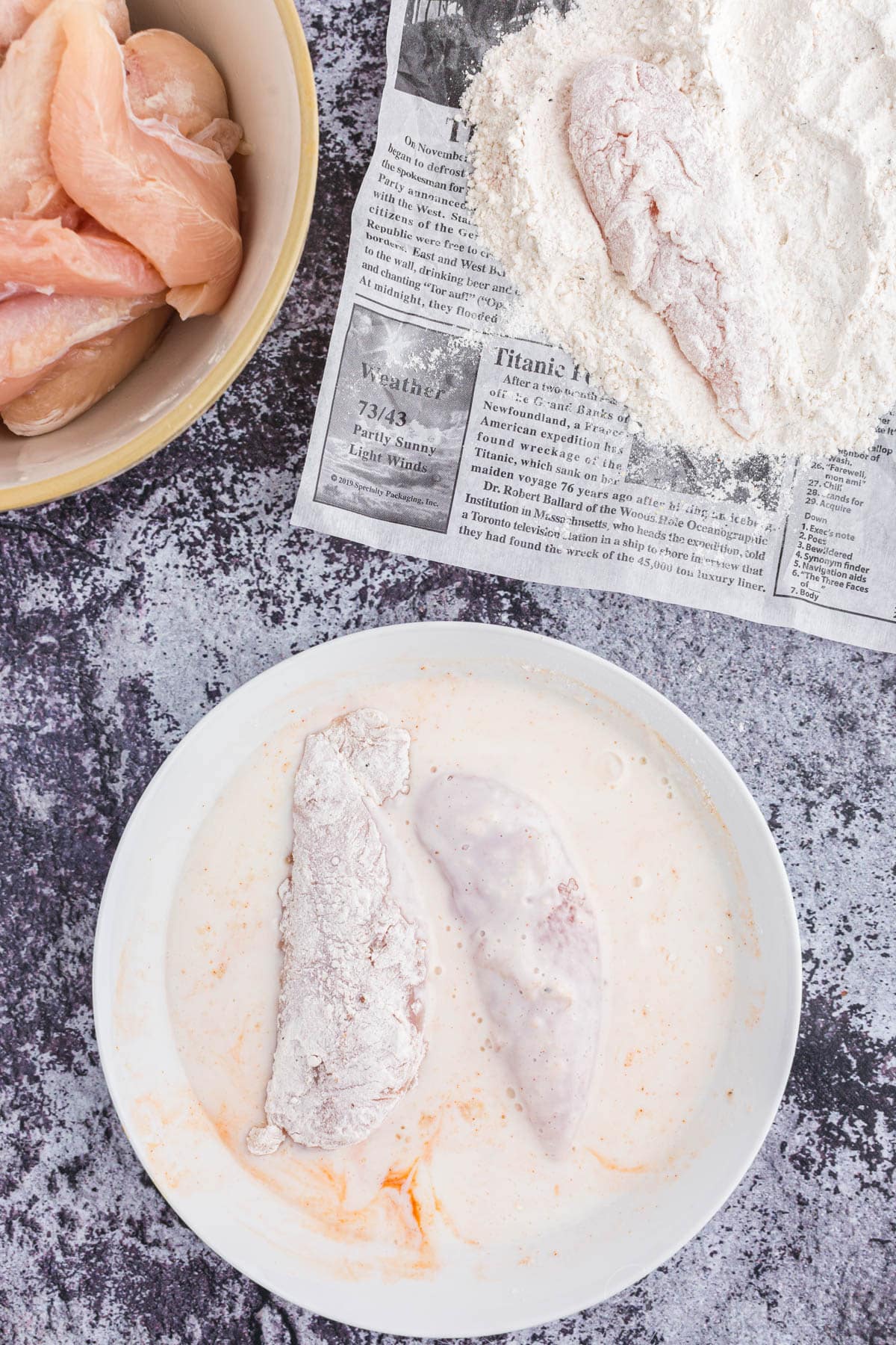 Overhead view of bowl with buttermilk mixture and flour-coated chicken tenders, bowl of raw chicken tenders, parchment paper with flour-coated chicken tenders.