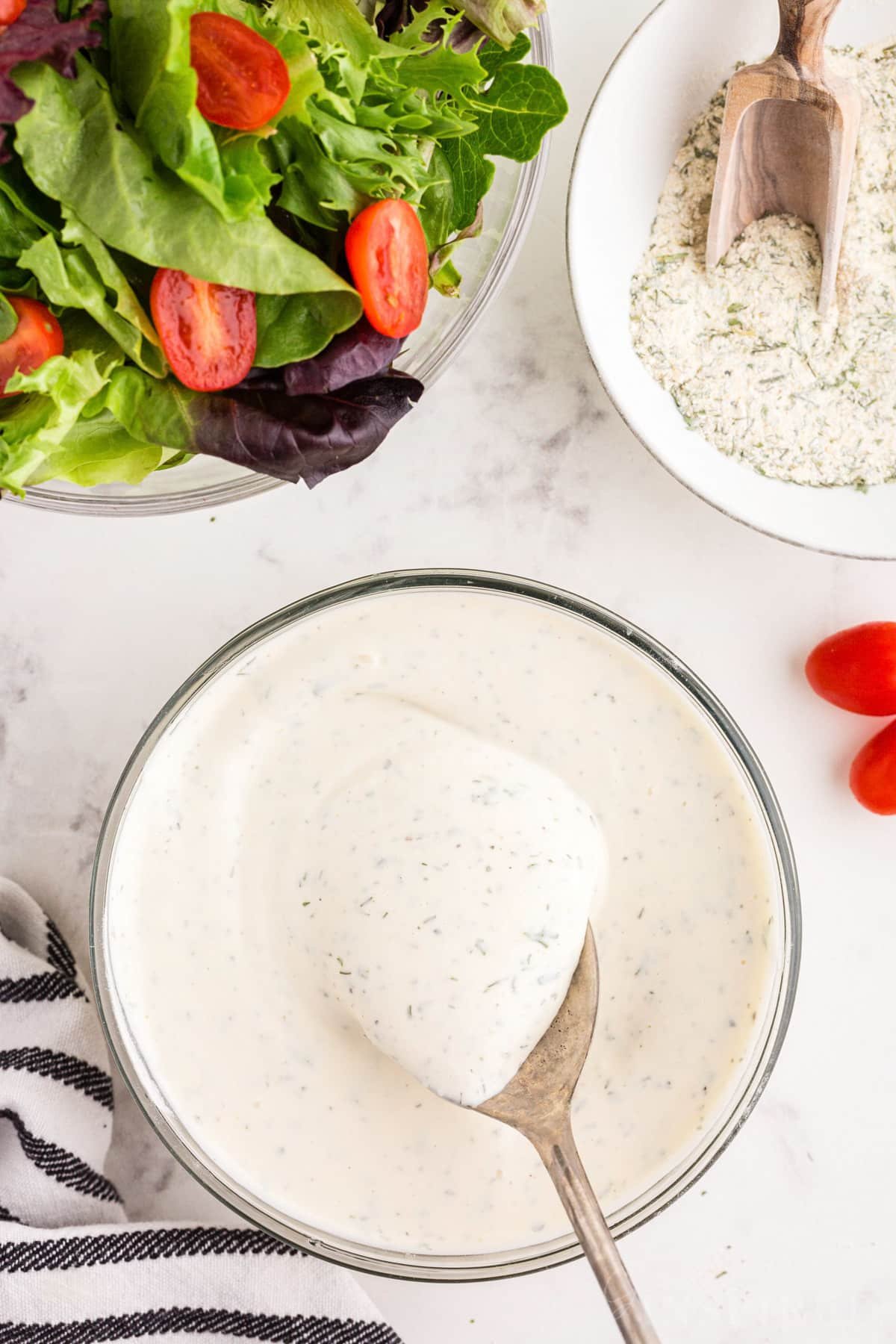 A bowl of Hidden Valley Ranch Dressing Recipe with a spoon inserted next to ranch seasoning and a salad.