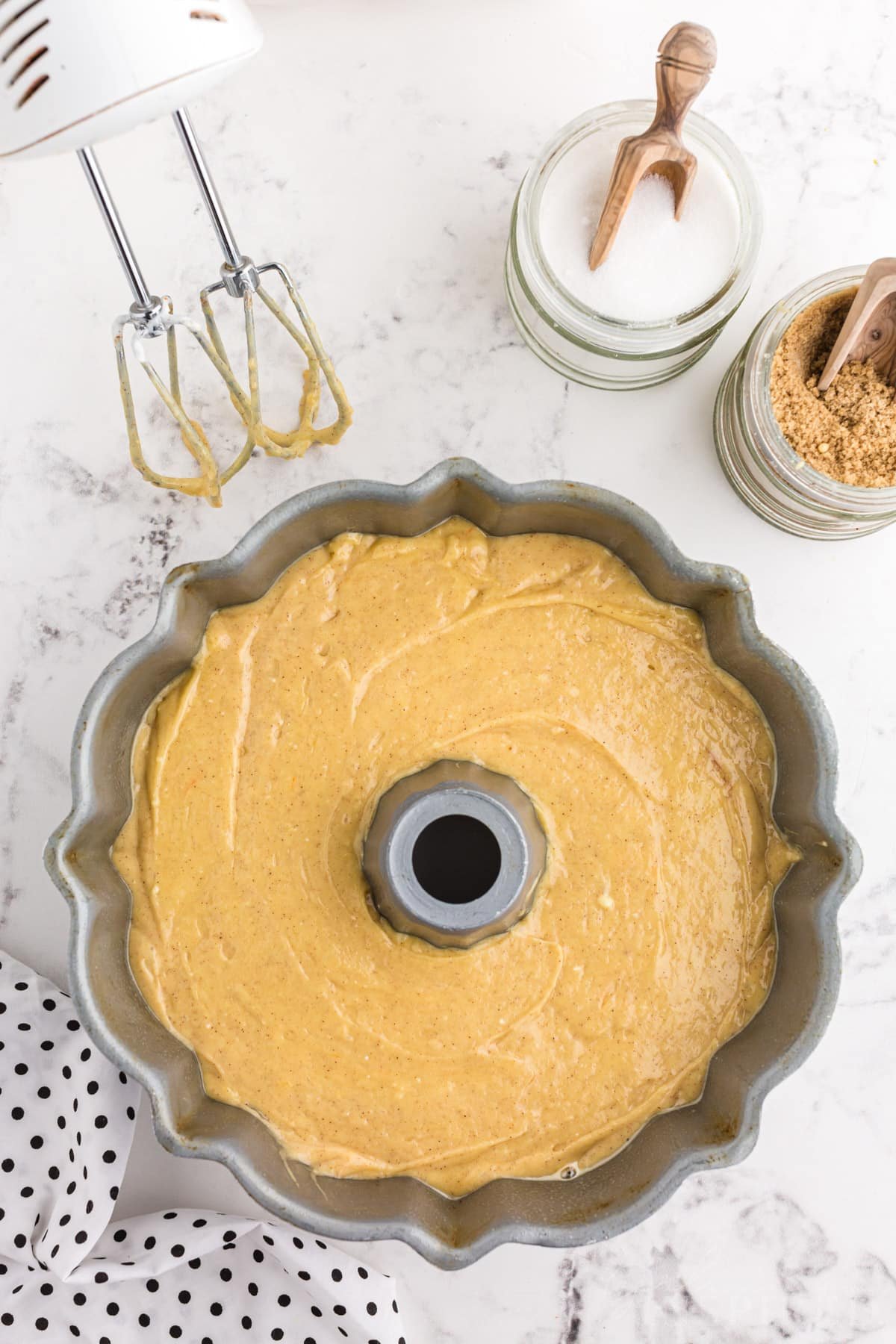 Crack cake batter poured into prepared bundt pan, hand mixer and cake ingredients placed around the pan.