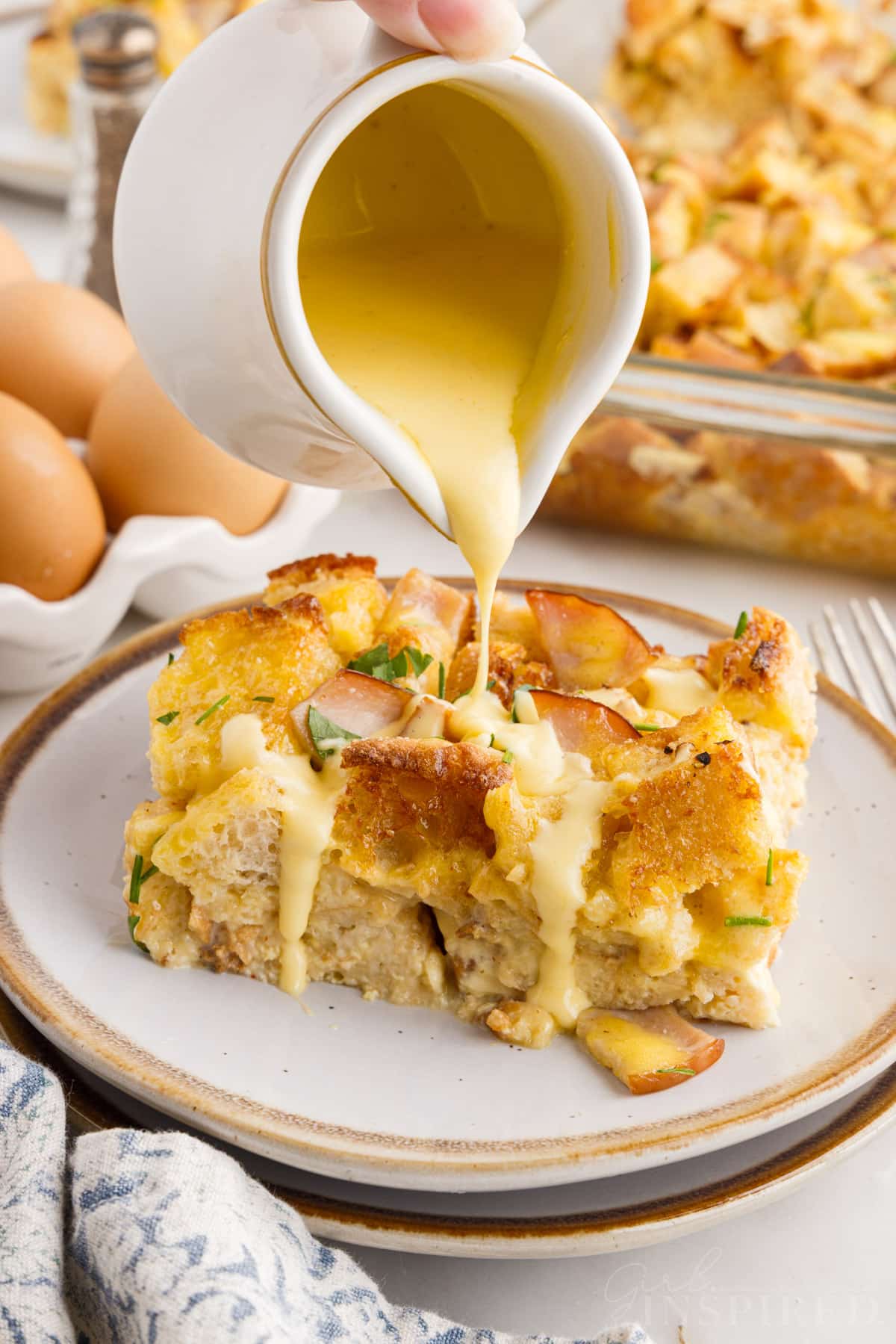 Hollandaise sauce poured over a slice of Eggs Benedict Casserole with the casserole in the background.