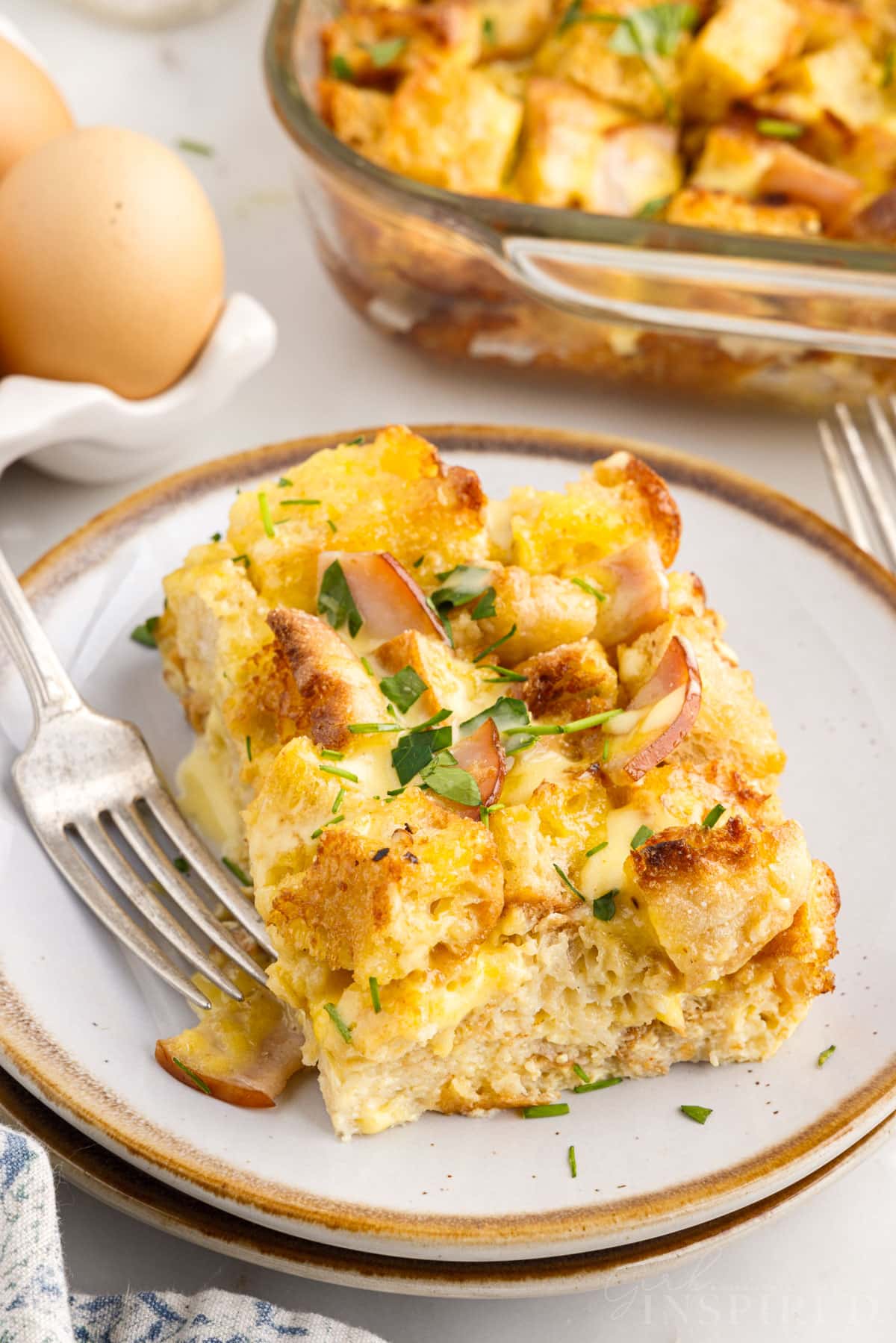 A dish with a slice of Eggs Benedict Casserole on it with a fork, eggs and casserole in the background.