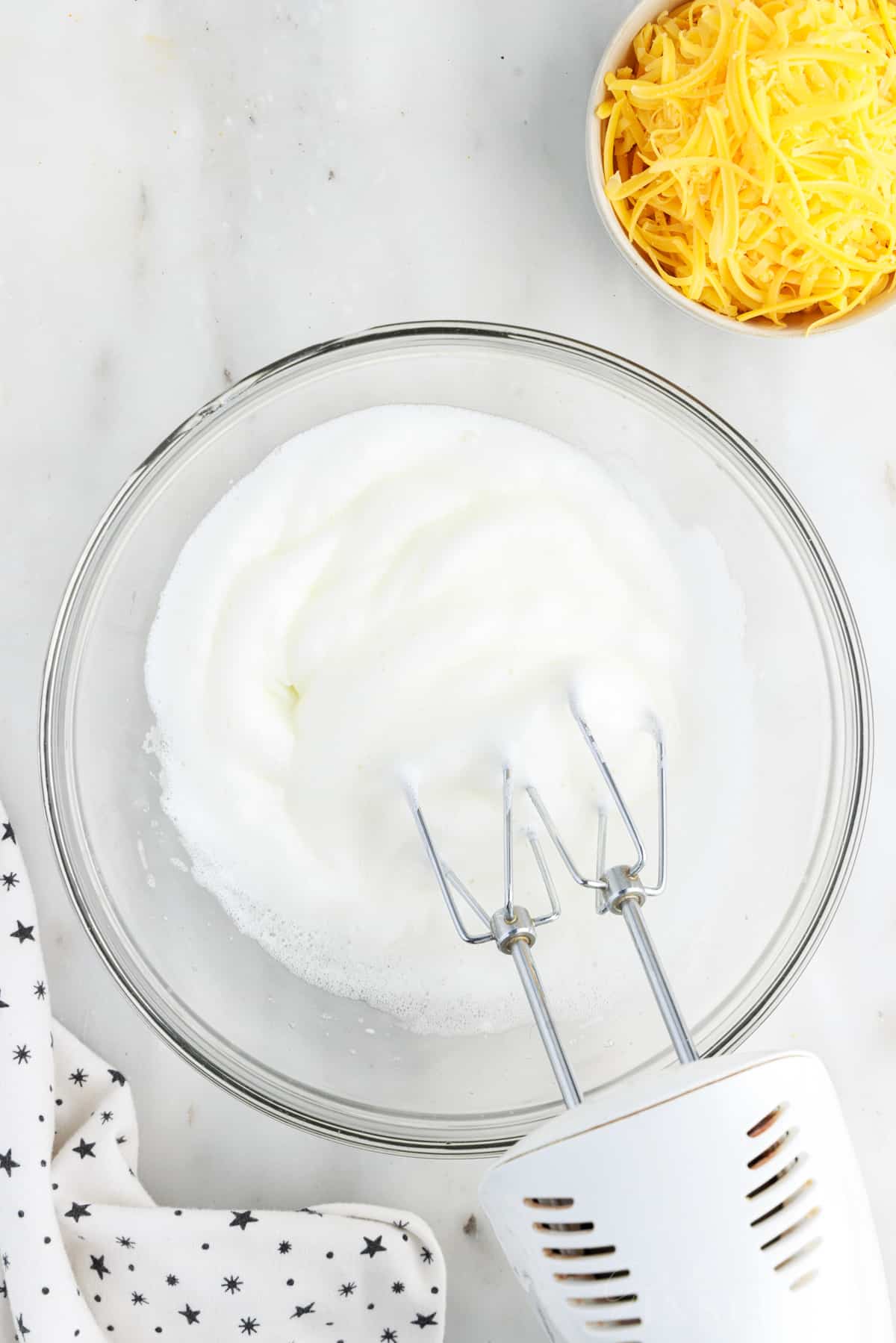 Stiff egg whites in a mixing bowl with mixer inserted next to a bowl of cheese.