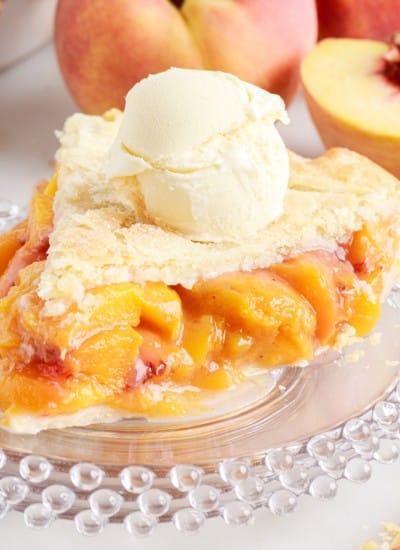Close up of a slice of peach pie on plates with peaches in the background.