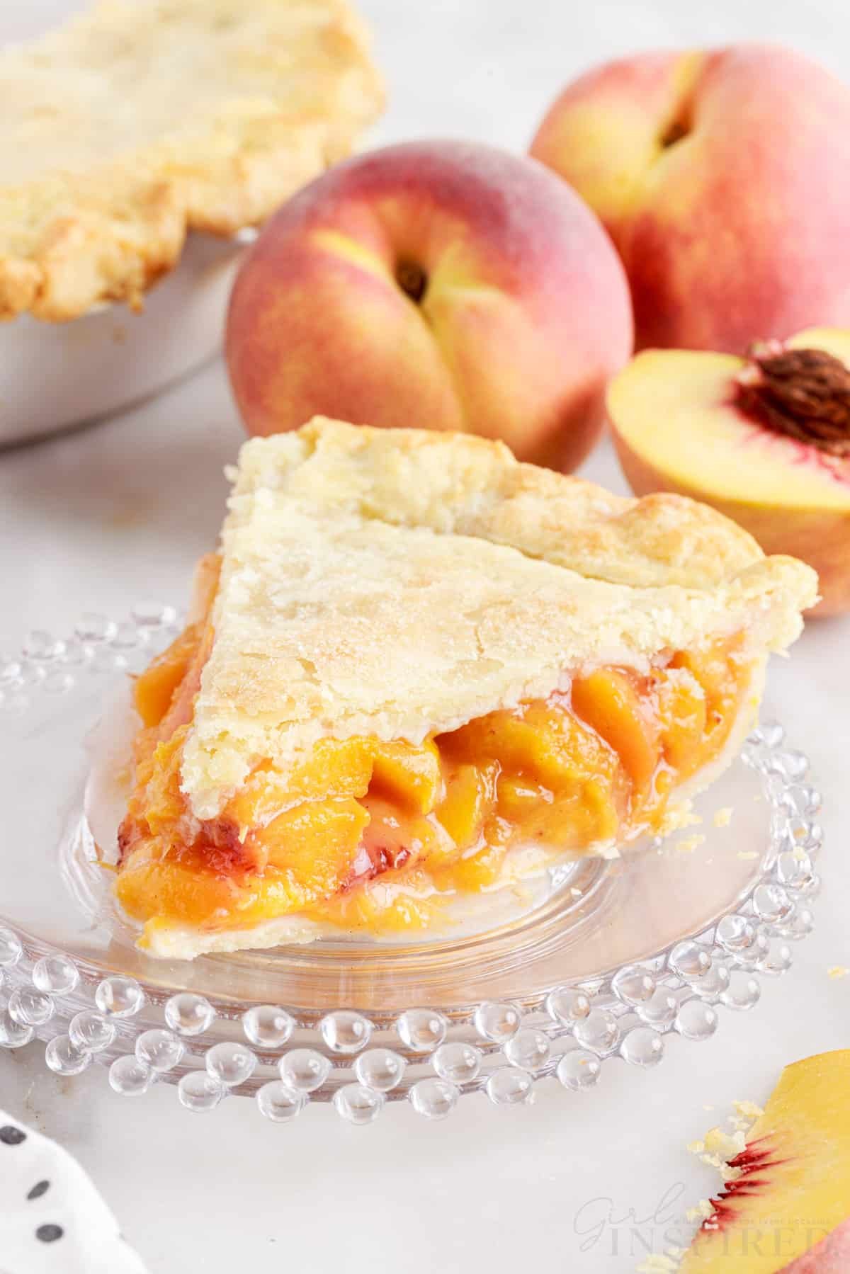 A slice of peach pie on a dish with peaches in the background.