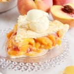 Stacked small plates with a slice of peach pie on it with ice cream, the pie in the background and peaches.