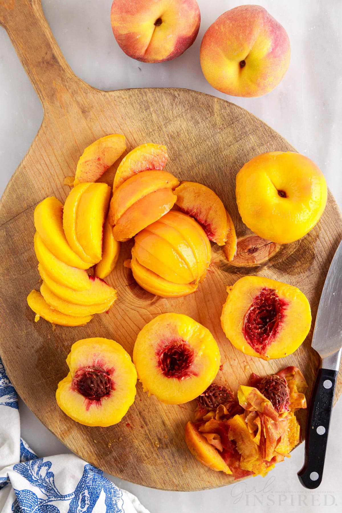 A round wooden cutting board with sliced peaches and halved peaches with a knife on the cutting board.
