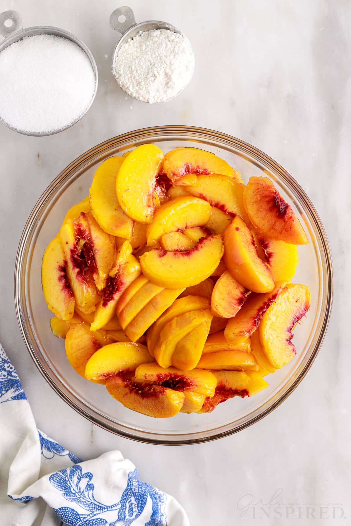 Sliced peaches in a glass mixing bowl next to a cup of sugar and flour.