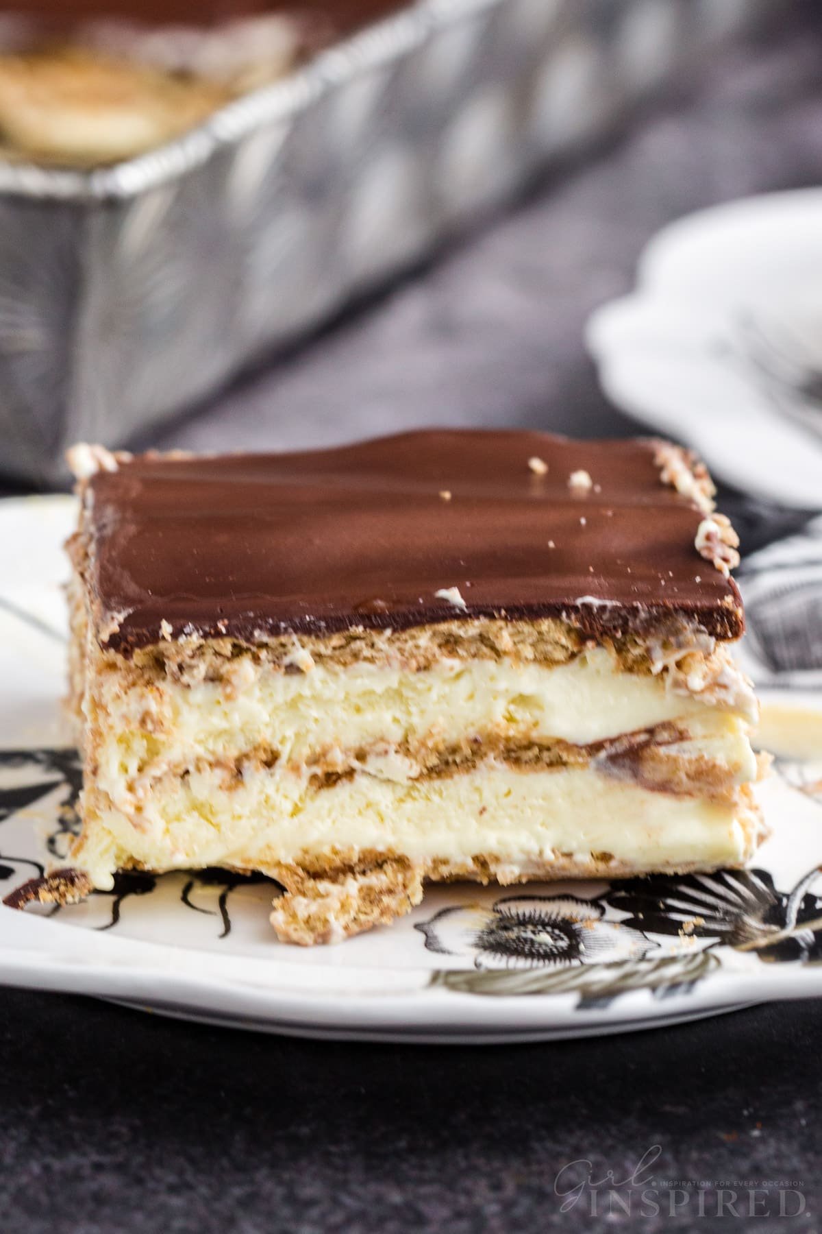 Single slice of no bake eclair cake on a serving plate, tray with remaining eclair cake in the background.