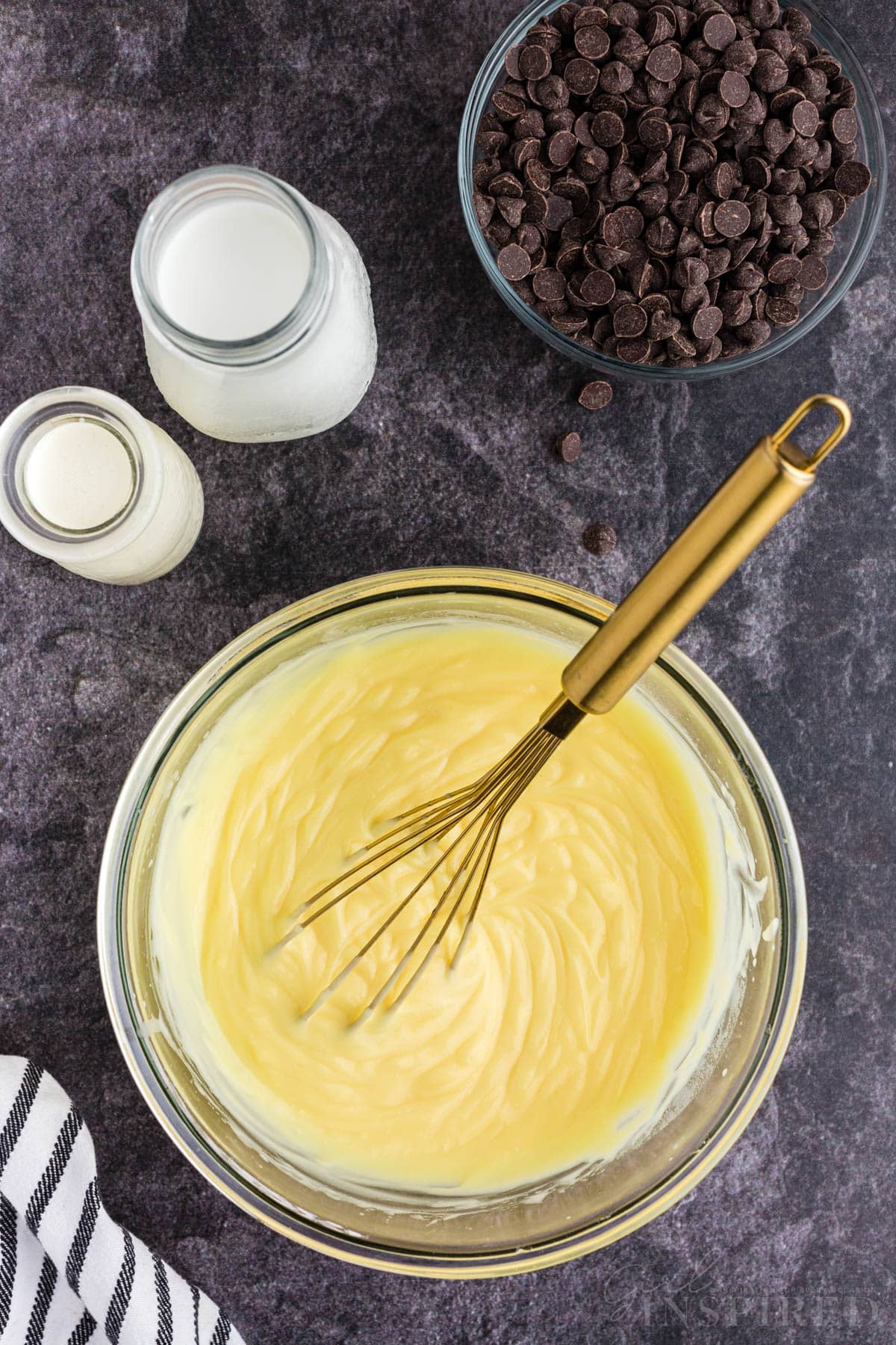 Mixing bowl with instant vanilla pudding mix and milk, metal whisk resting the bowl, no bake eclair cake recipe ingredients around the mixing bowl.
