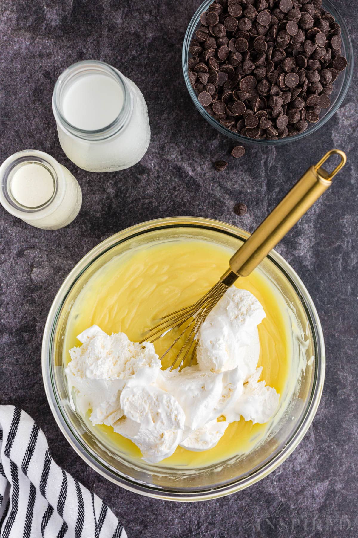 Mixing bowl with vanilla instant pudding, milk, and Cool Whip, metal whisk in the bowl, no bake eclair cake recipe ingredients on the countertop.