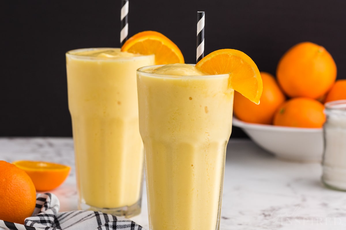 Two glasses of Orange Julius recipe with decorative straws, bowl of fresh oranges in the background.