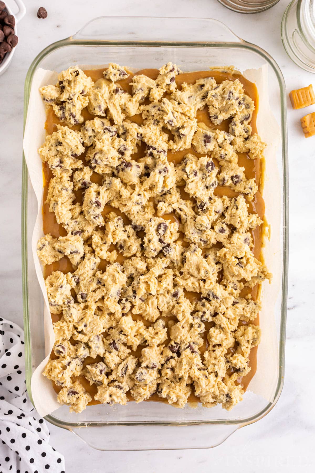 Overhead view of salted caramel chocolate chip cookie bar mixture in a prepared baking pan with freshly crumbled cookie dough topping.