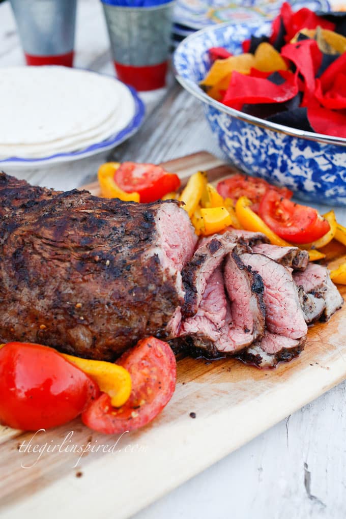 marinated tri tip on wooden cutting board with veggies, paper plates and utensils, bowl of red and blue tortilla chips.