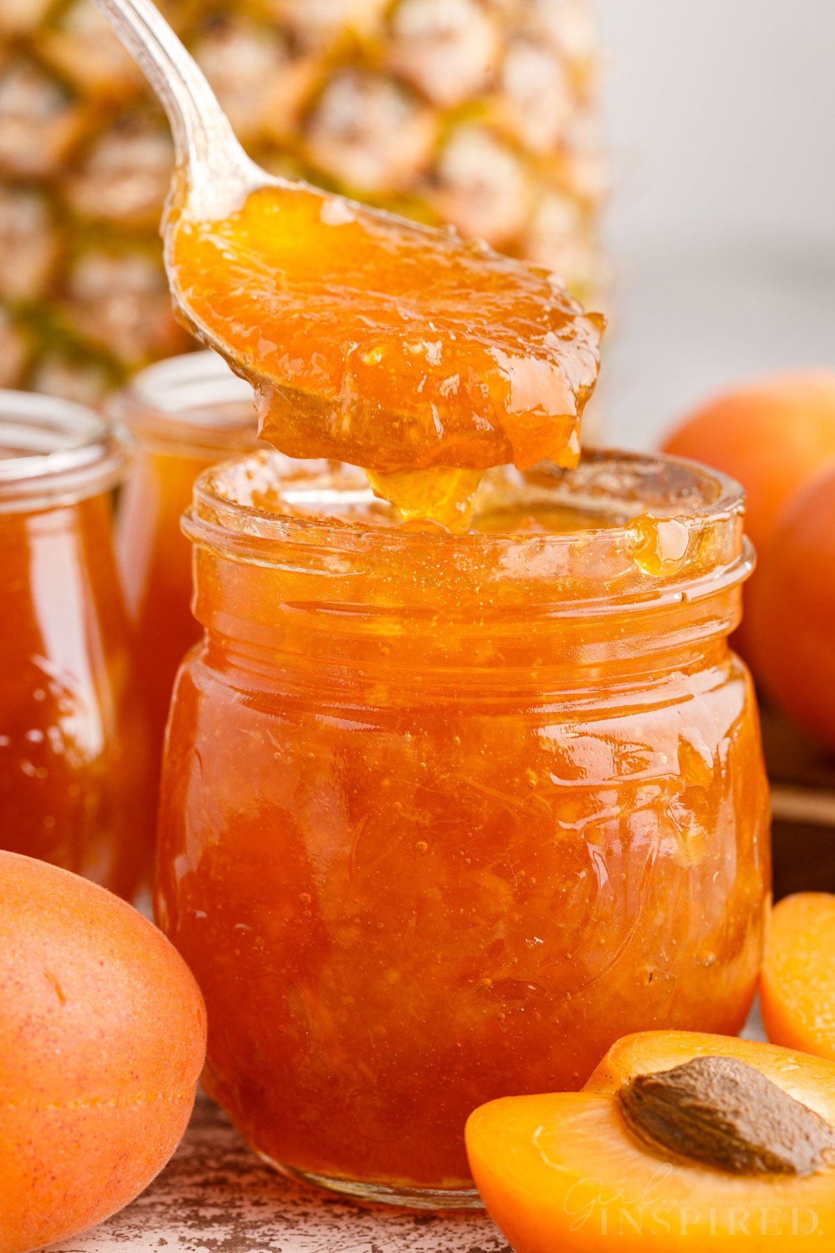 Apricot Pineapple Jam in a glass jar with a spoonful of jam, fresh apricots and pineapple in the background.