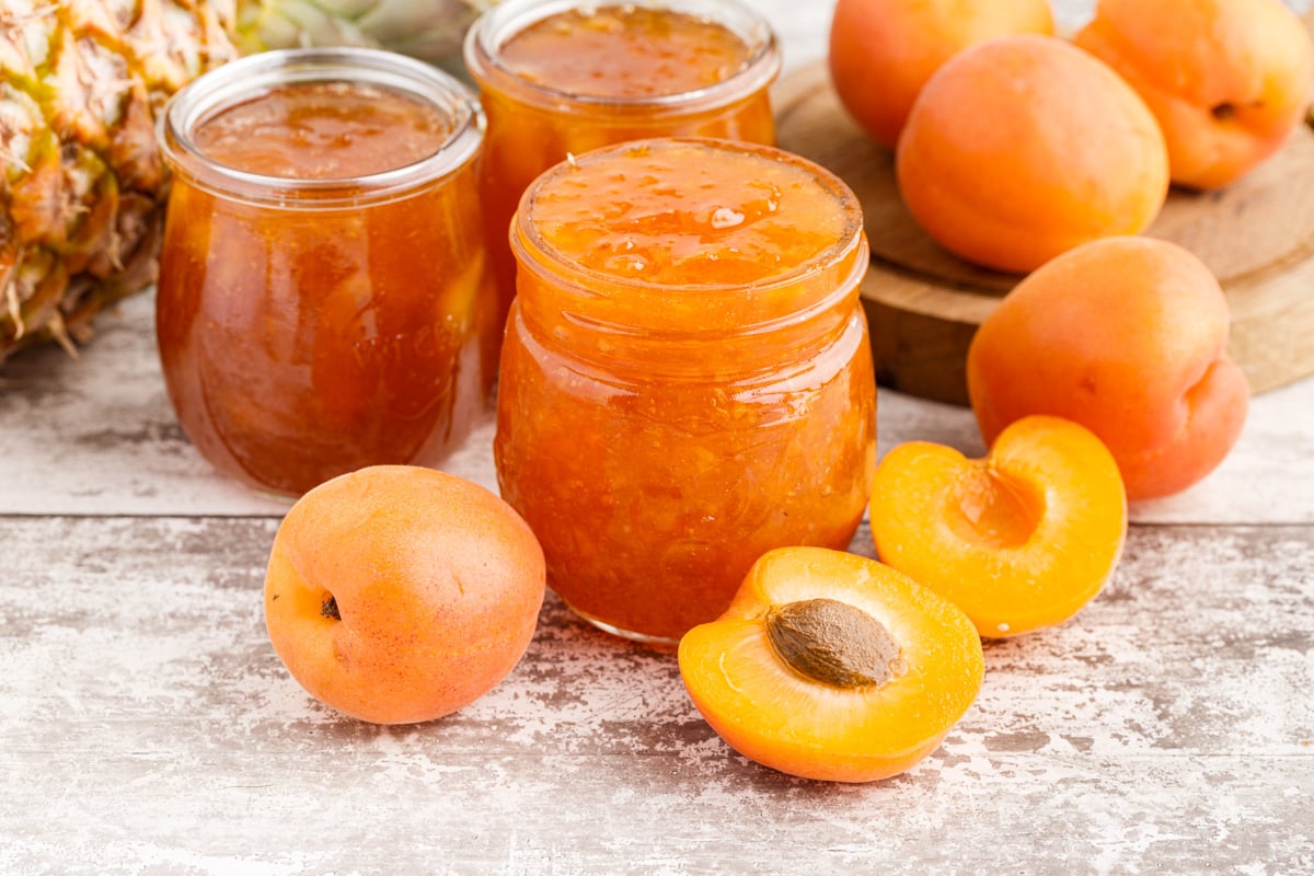 Glass jars filled with Apricot Pineapple Jam, fresh apricots and pineapple on a wooden countertop.
