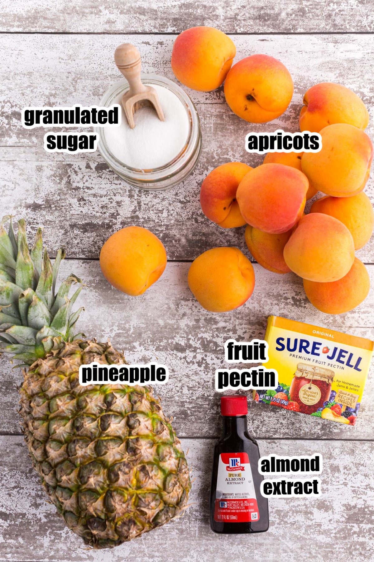 Individual ingredients (pineapple, apricots, sugar in a bowl, pectin in a box, almond extract in a bottle) needed for making apricot pineapple jam with text labels.