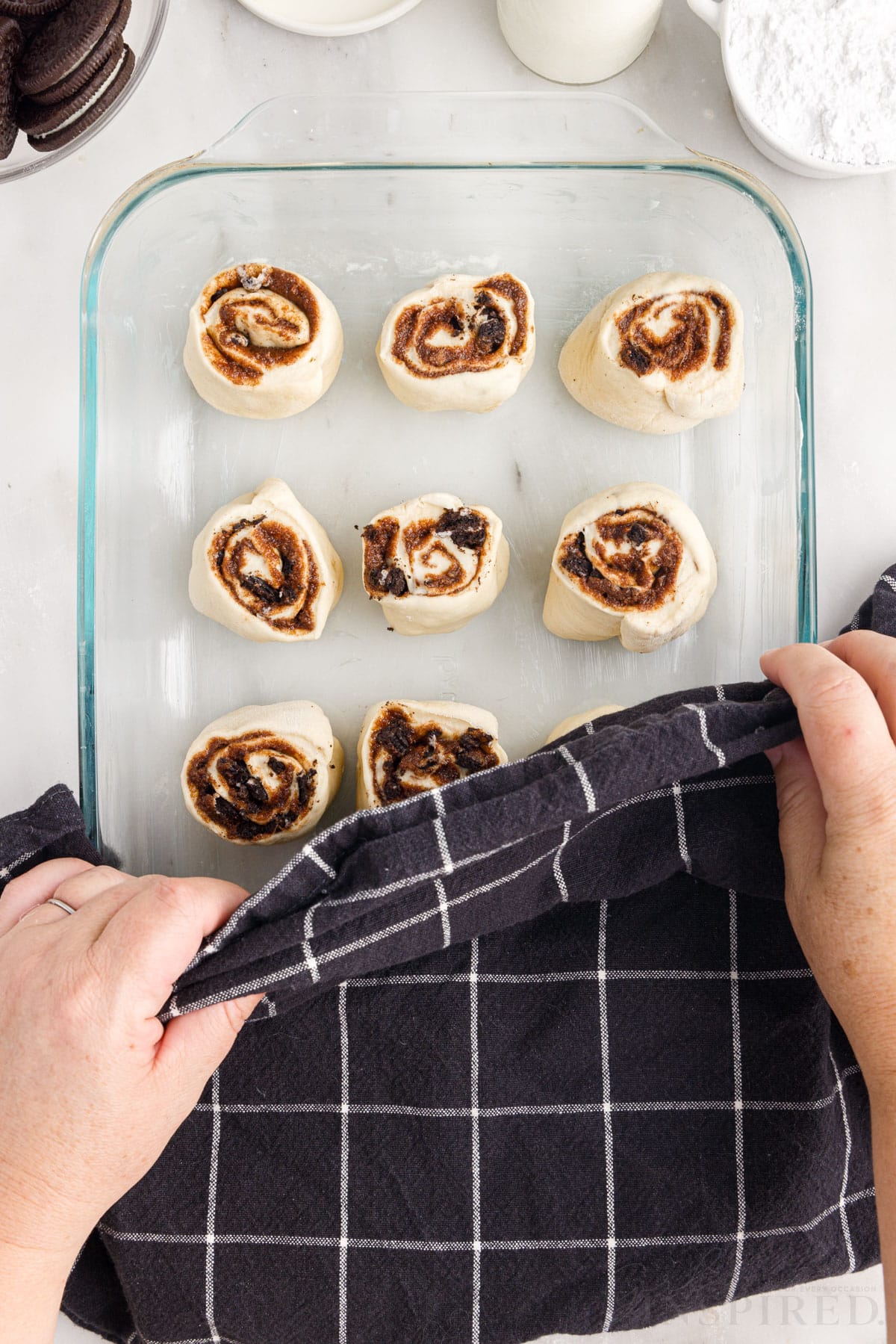 Cookies and Cream Cinnamon Rolls in glass 9x13 pan before baking being covered with a towel.