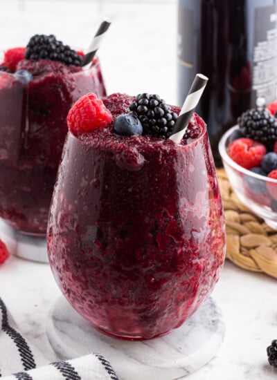 Two Frozen Wine Slushies in glasses garnished with raspberries, blueberries, and blackberries. A bowl of berries in the background.