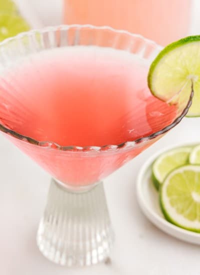 Decorative glass with Pink Flamingo Drink garnished with a lime next to a dish of limes.