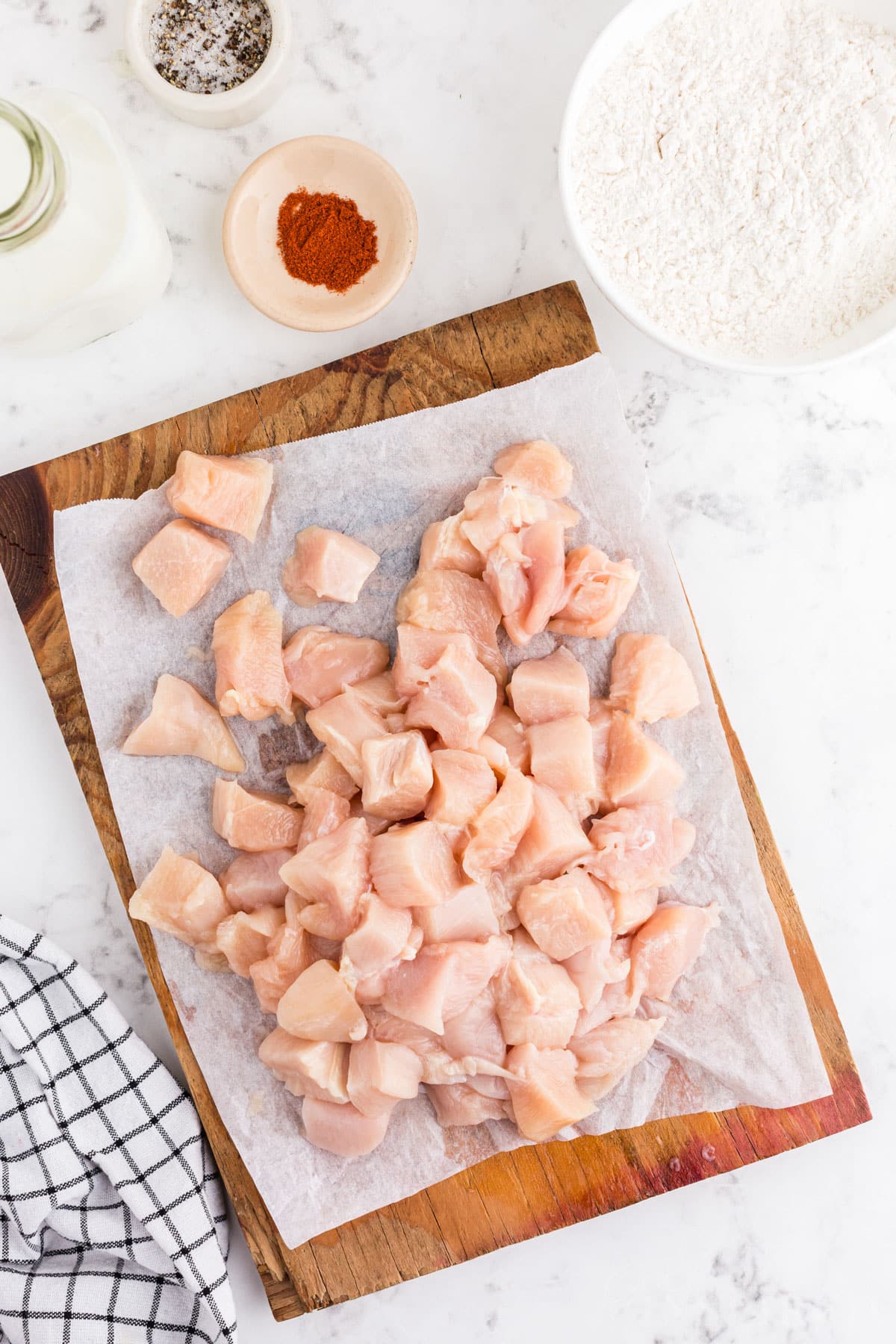 Diced chicken pieces on parchment-lined wooden kitchen board, dredging ingredients surrounding the chicken.