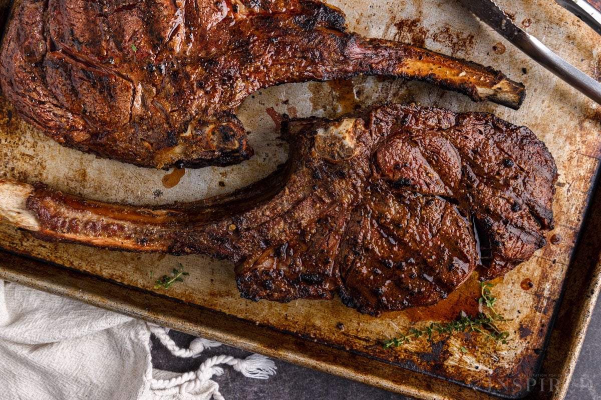 Two cooked Tomahawk steaks on a metal baking sheet with metal tongs.