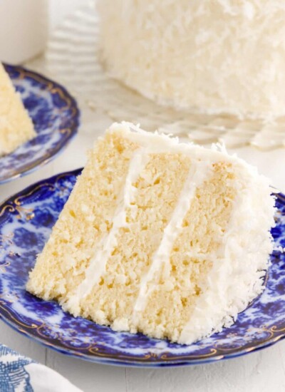 A slice of Coconut Cream Cake on a small dish with the cake in the background.