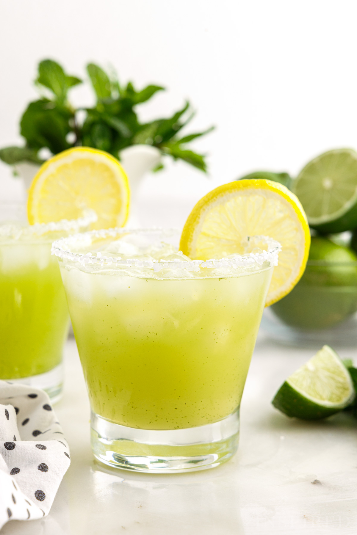 Two short glasses of Mint Margarita garnished with lemons, limes in the background.
