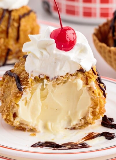 Deep fried ice cream with a bite taken out on a plate topped with whipped cream and a cherry.