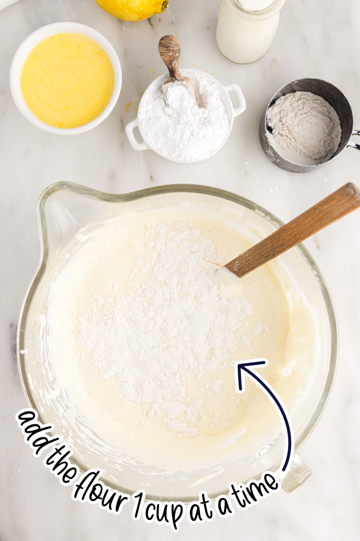 Flour added to the egg mixture in a mixing bowl with a spatula inserted with text overlay.