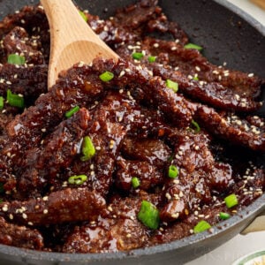 Skillet full of ginger beef garnished with green onions and sesame seeds, with wooden spoon scooping.