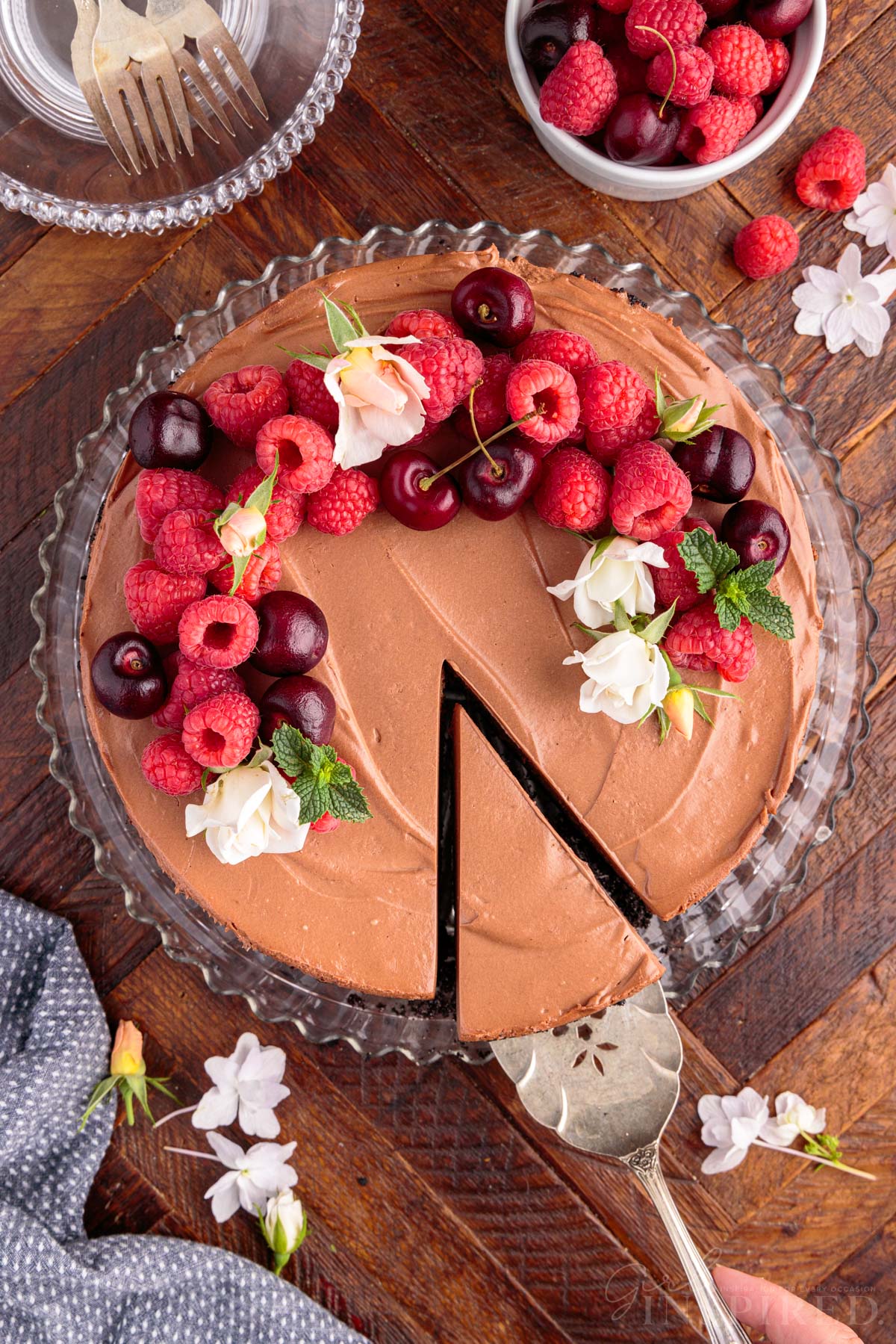 No Bake Chocolate Cheesecake garnished with flowers, cherries, raspberries, and mint leaves, with pie server lifting the first slice.
