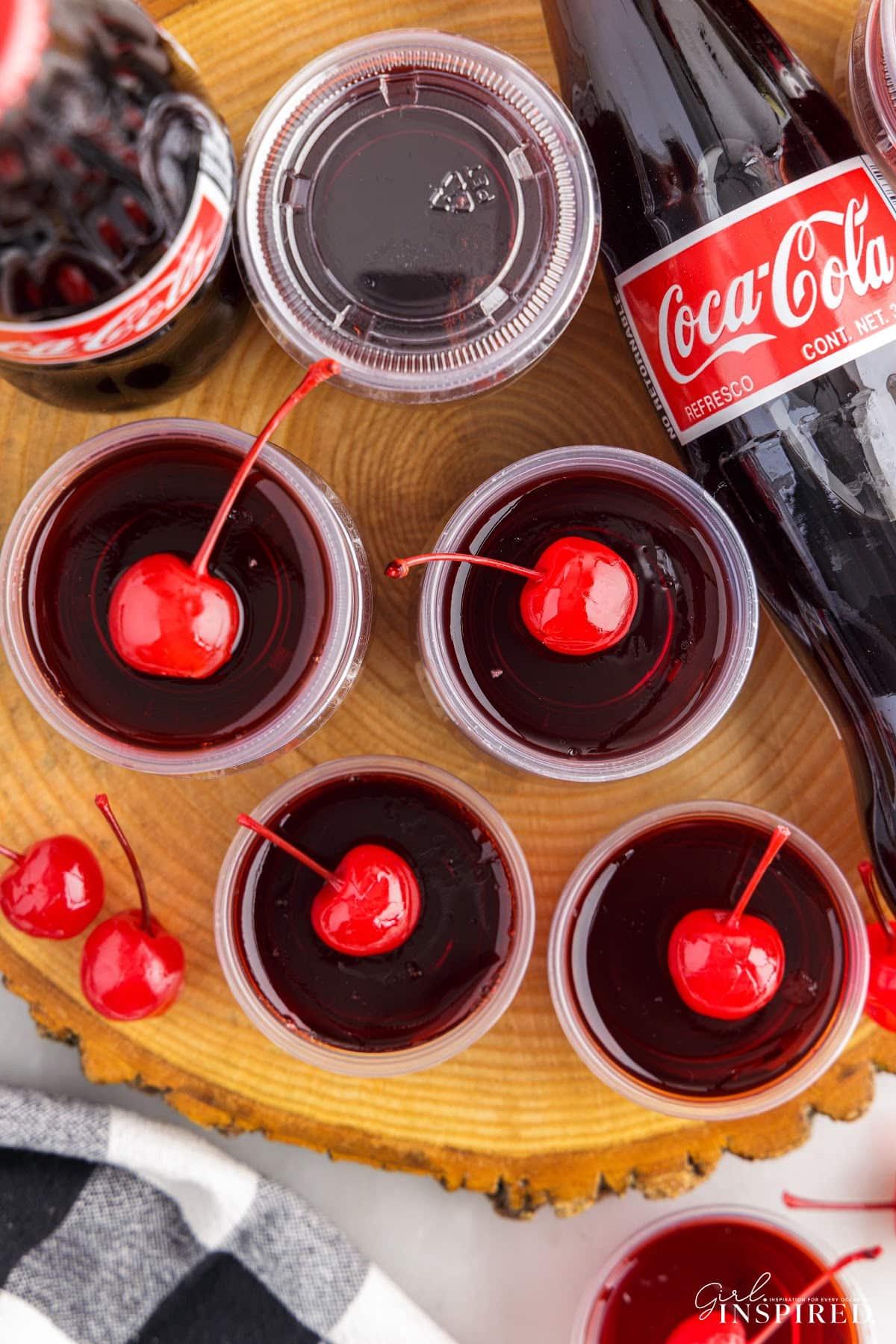 Cherry Coke Jello Shots topped with cherries, on wooden round, glass bottle of Coca-Cola.