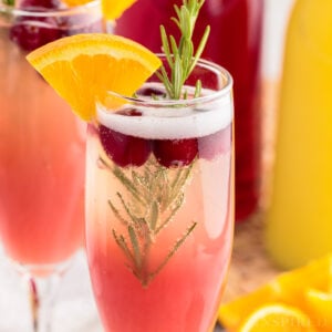 Two Cranberry Orange Mimosas garnished with orange wedge and rosemary sprig next to carafes of juice.