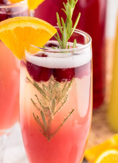 Two Cranberry Orange Mimosas garnished with orange wedge and rosemary sprig next to carafes of juice.