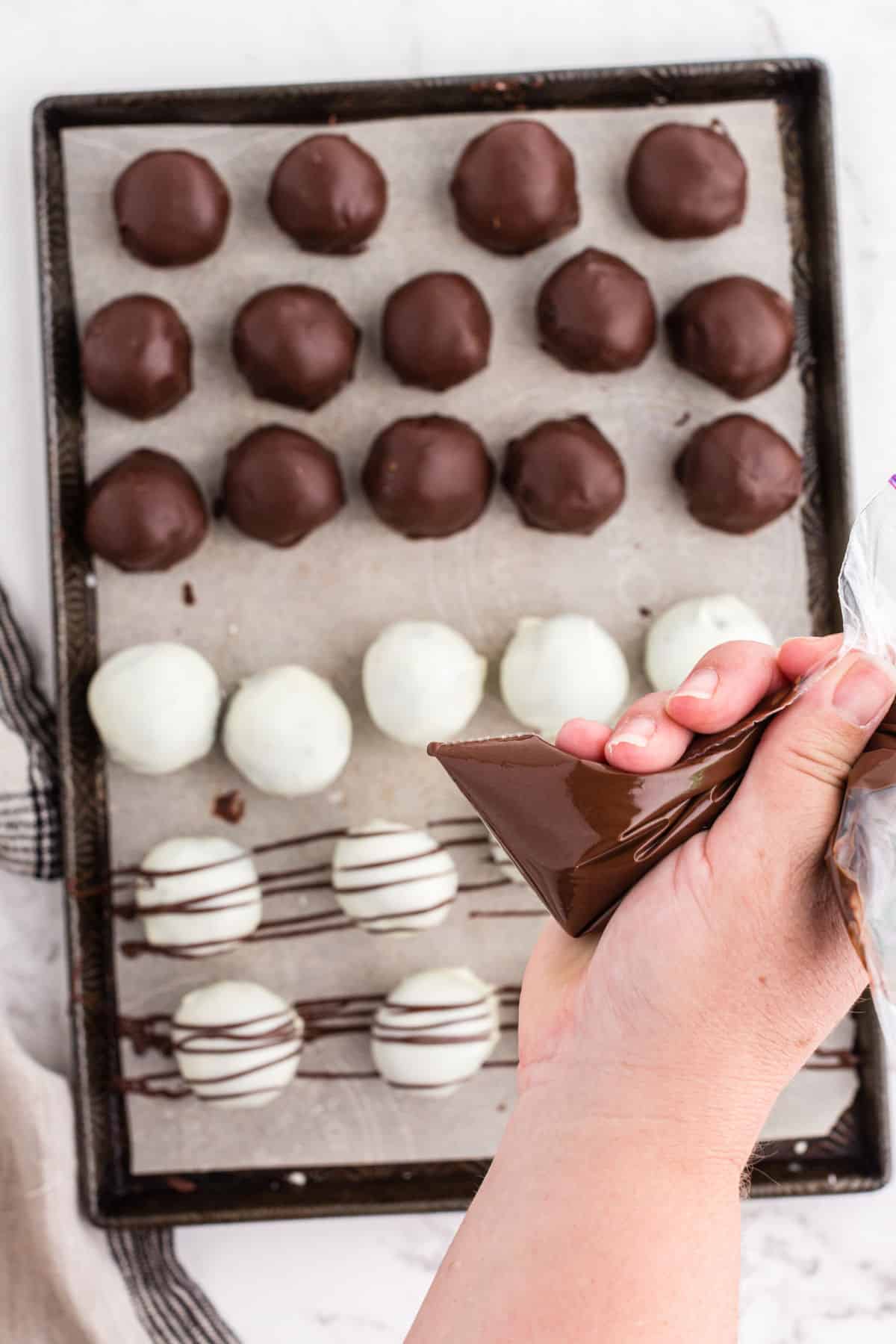 Holding a plastic baggy with melted chocolate over tray of Oreo balls and drizzling the chocolate over the balls.