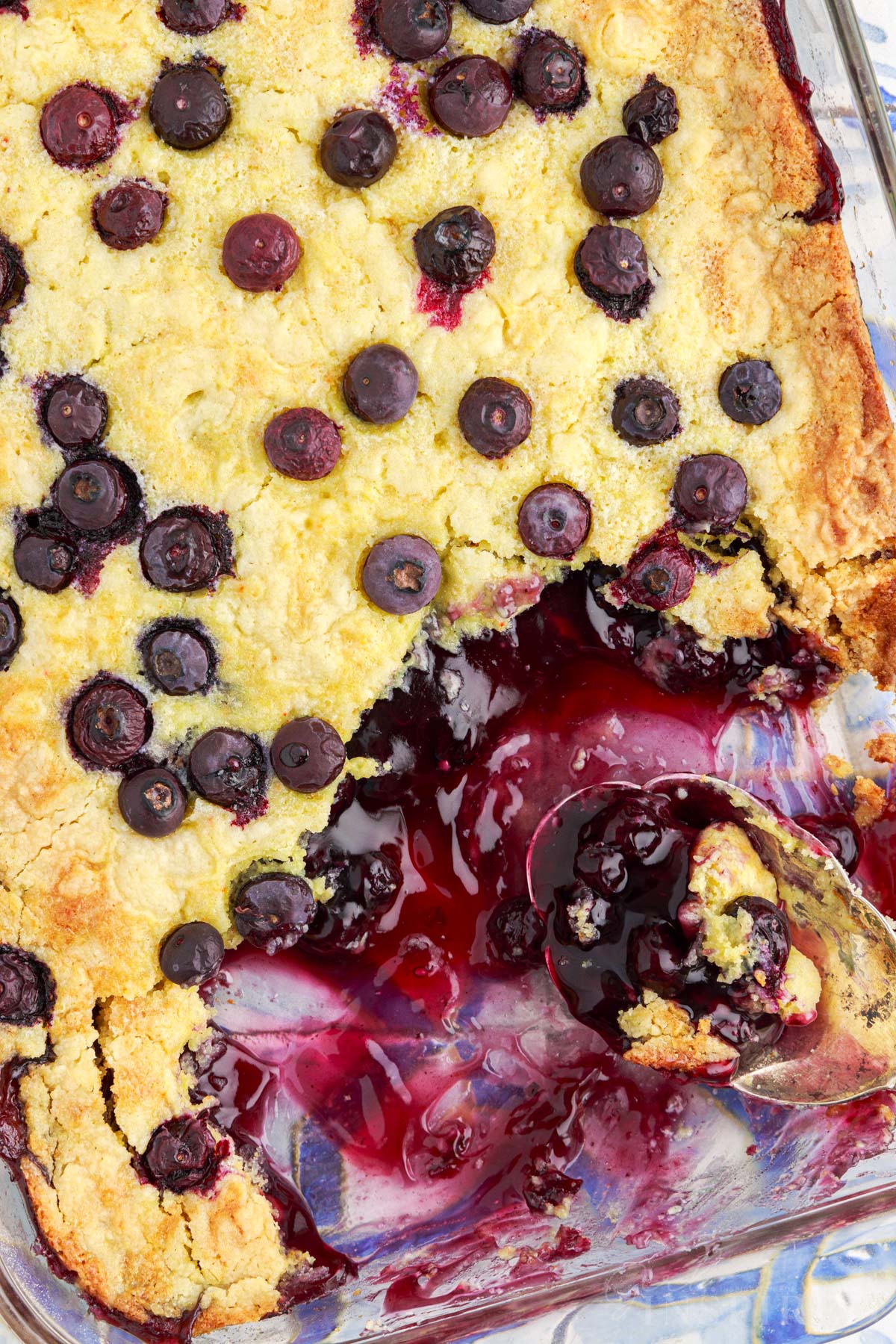 Overhead view of Blueberry Dump Cake in glass casserole dish with a spoon laying in pan where a scoop has been removed, with text title overlay.