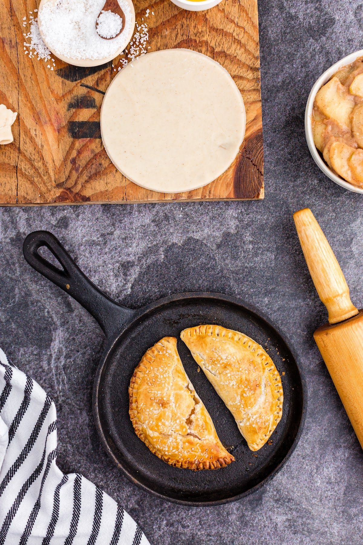 Two fully cooked hand pies in a cast iron skillet, with rolling pin and cutting board with pie crust.
