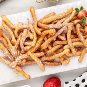 A platter of Funnel Cake Fries sprinkled with powdered sugar, with fresh strawberries as garnish.