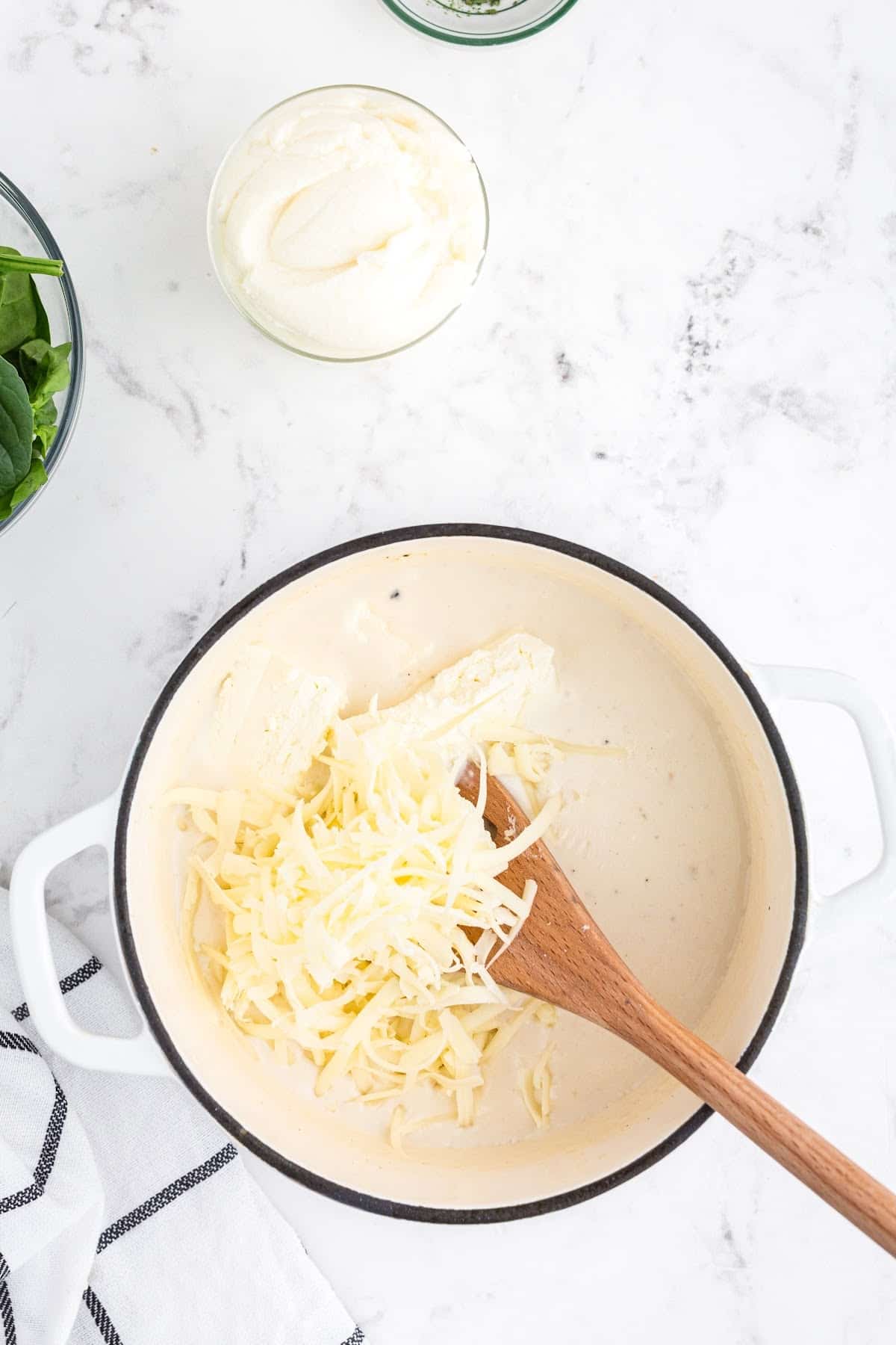 Cheese and cream cheese being added to the mixture in the dutch oven. Smaller bowls on the side of spinach and cream cheese.