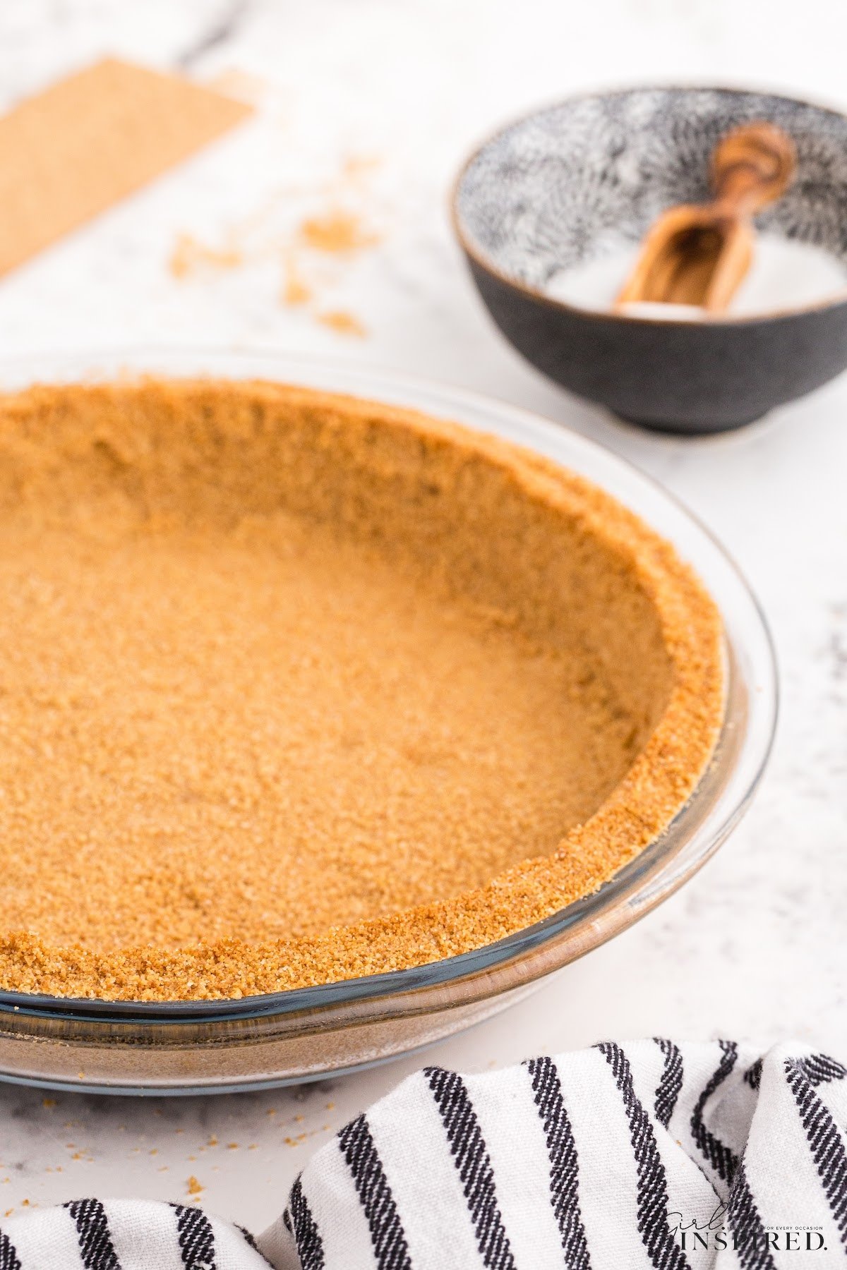 A graham cracker pie crust after setting in a glass pie plate next to a towel and sugar.