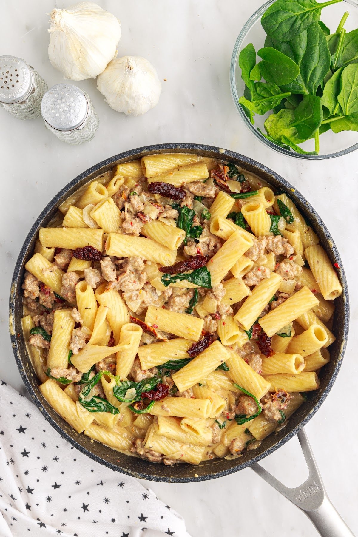 Creamy Italian sausage pasta in a large skillet next to garlic and spinach.