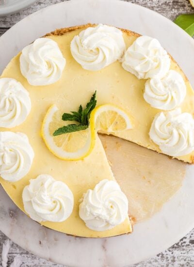 A lemon cheesecake on a serving tray with a slice taken out garnished with whipped cream and fresh lemon.