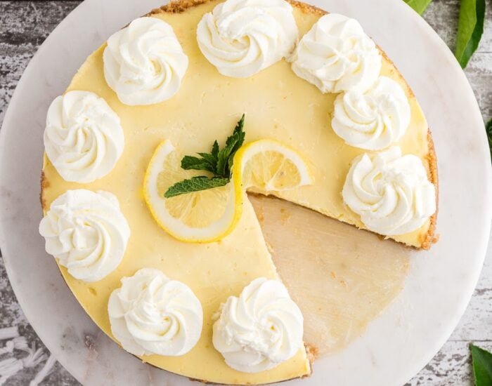 A lemon cheesecake on a serving tray with a slice taken out garnished with whipped cream and fresh lemon.