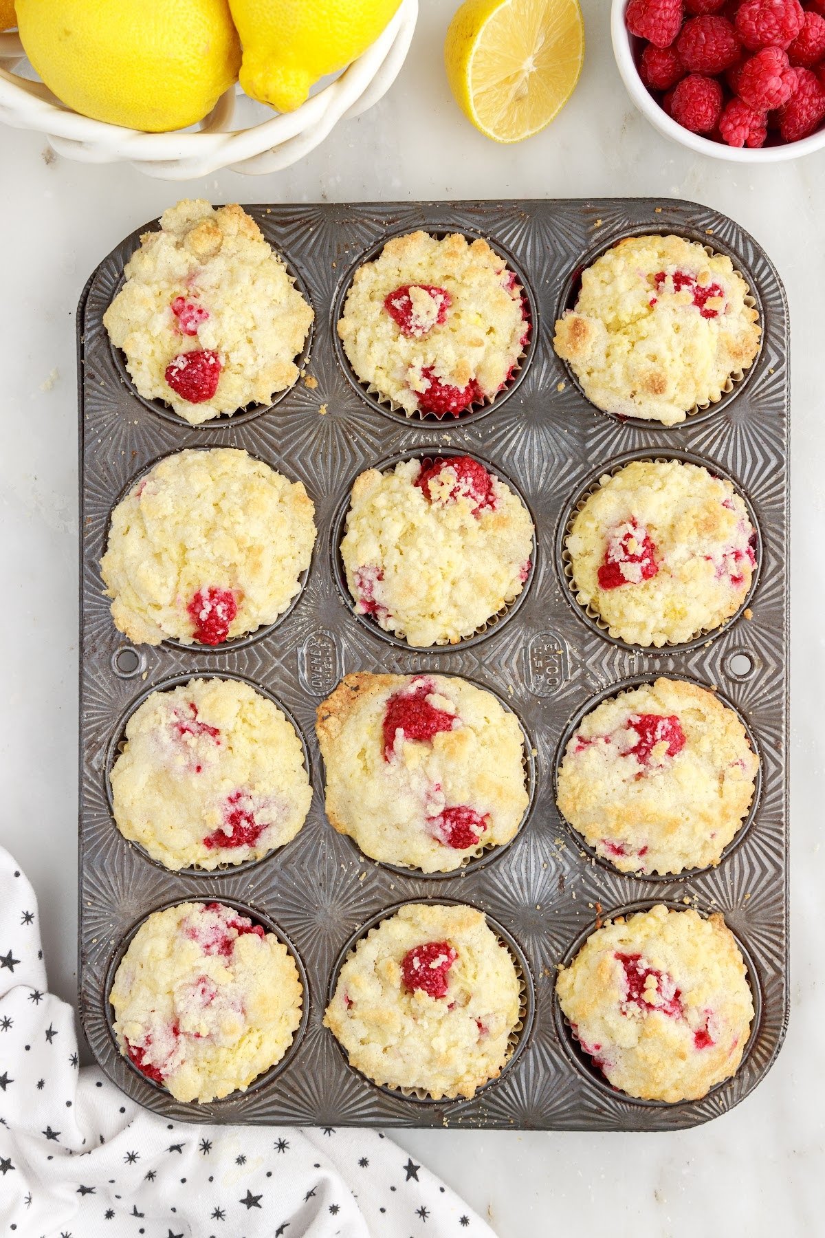 Baked lemon raspberry muffins in muffin tin next to bowl of lemons and bowl of raspberries.