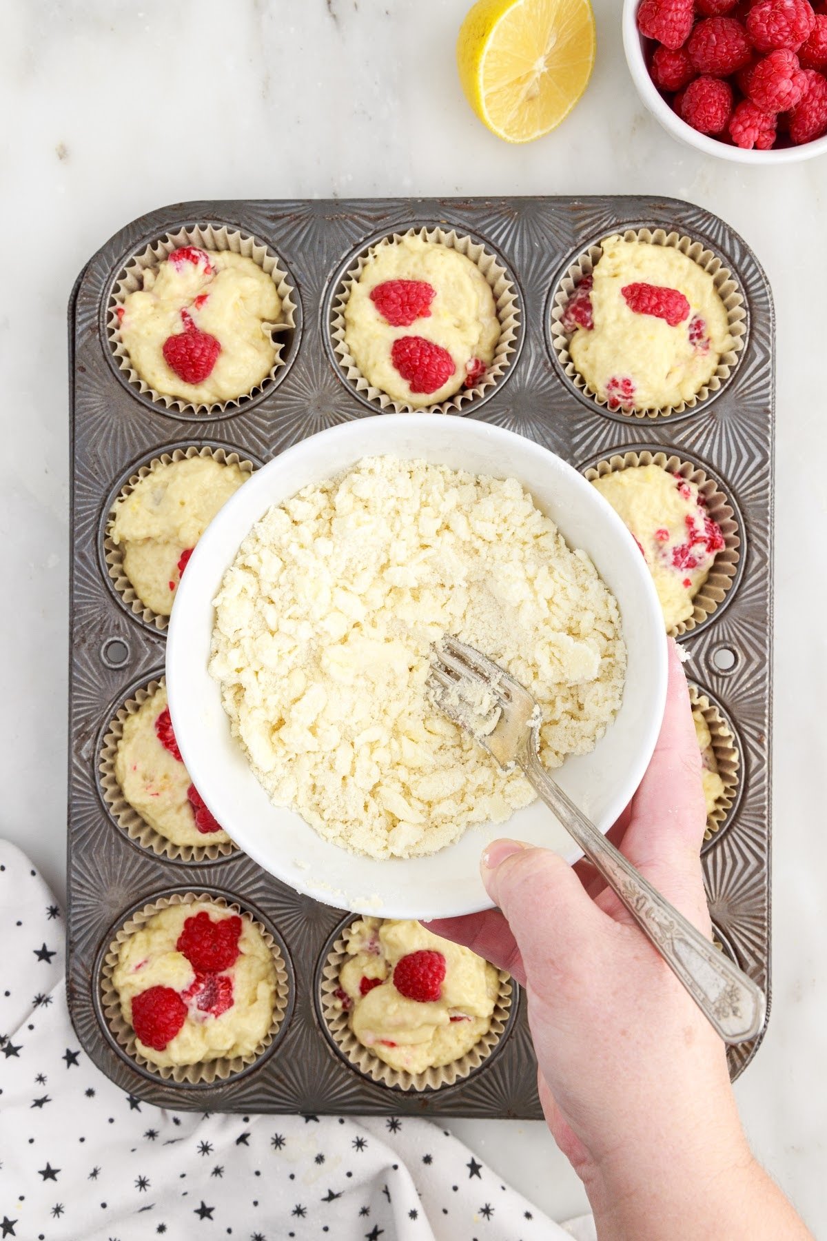 Streusel in a mixing bowl with a fork held over tray of raspberry lemon muffin batter.