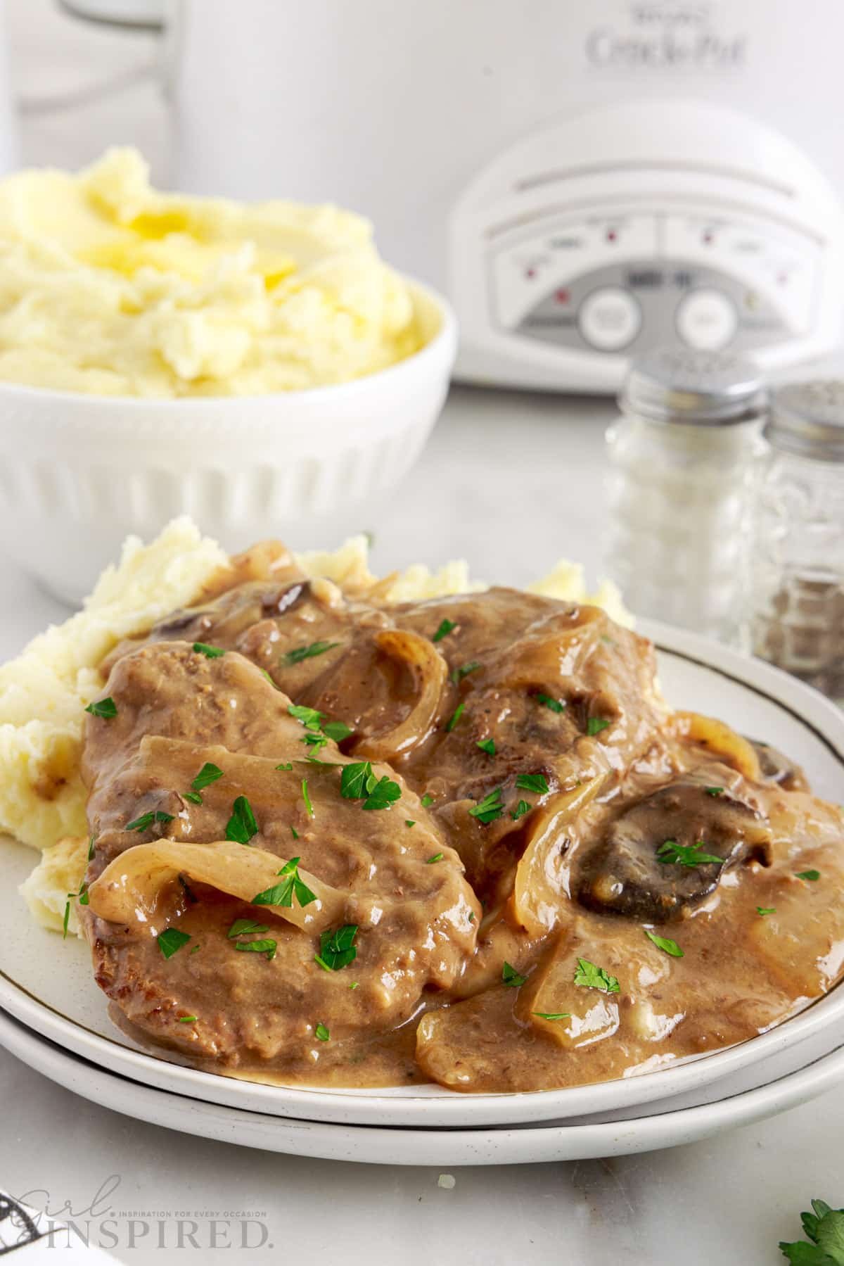 Crock pot cube steak smothered in onions and gravy, plated on a bed of mashed potatoes.