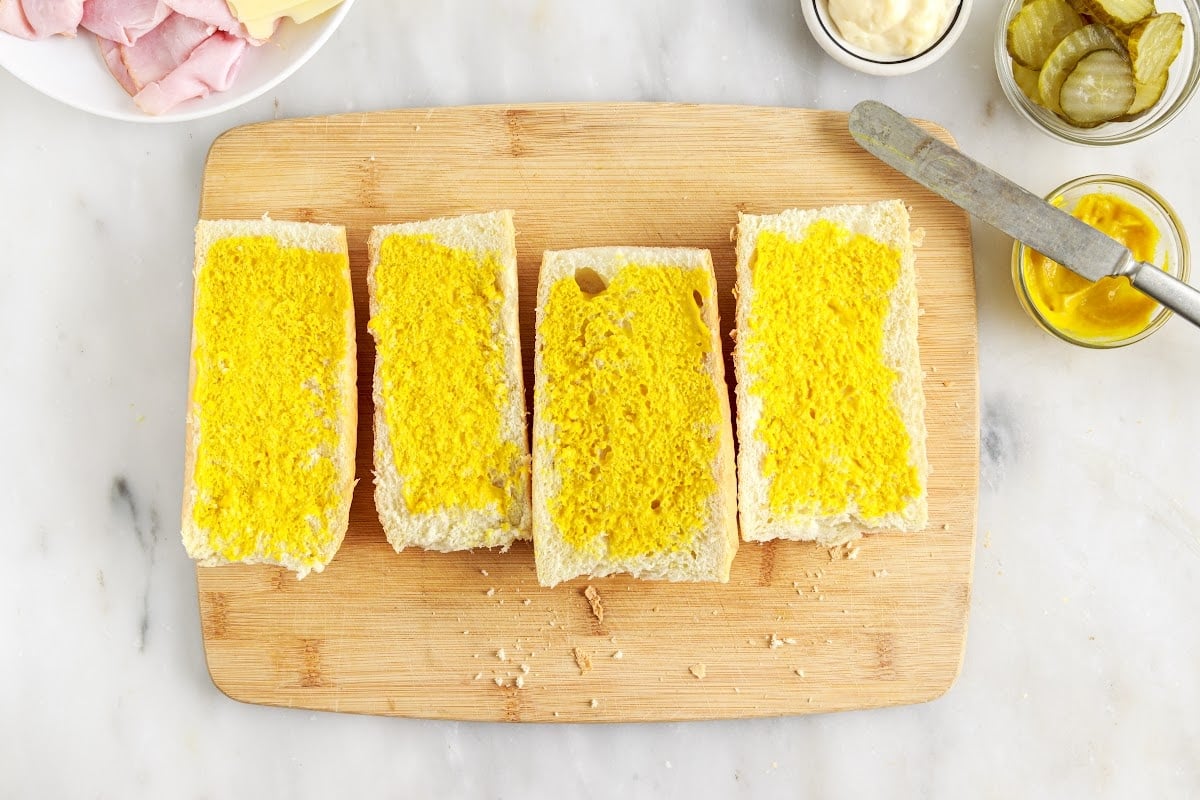 Sliced bread open faced on a cutting board, covered with mayonnaise and mustard.