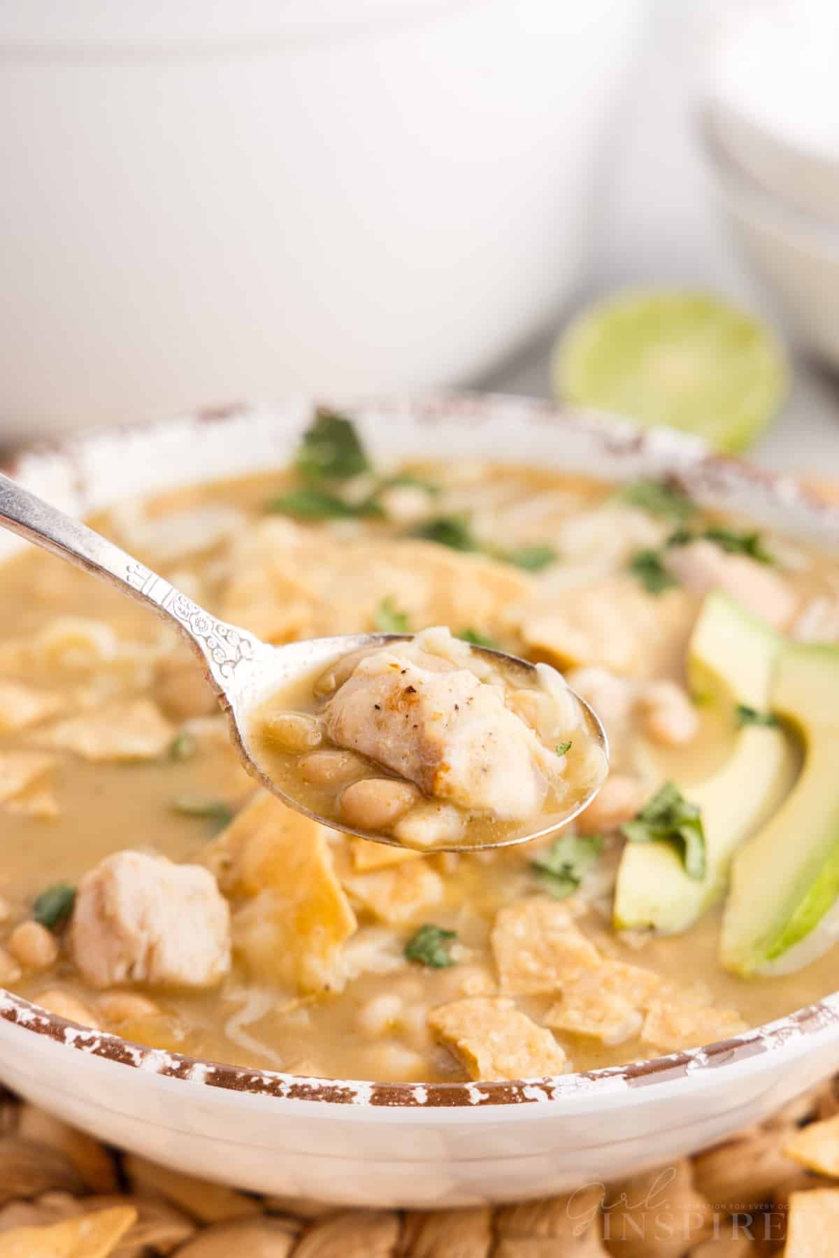 Spoon lifting chunk of chicken and beans from bowl of green chicken chili.