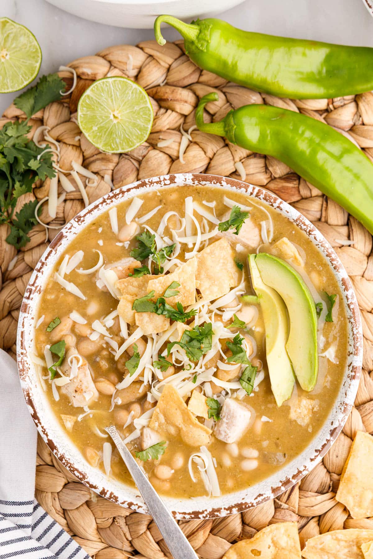 Bowl of green chicken chili garnished with cilantro, avocado slices, melted cheese, on a woven mat with green chiles and lime halves.